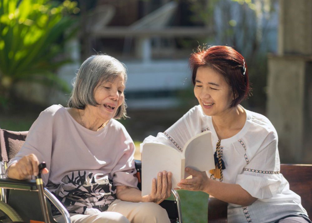 Older Asian Woman reading book with carer as part of her aged care package